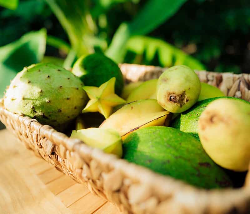 A wicker basket filled with various tropical fruits, including starfruits, mangoes, and other green fruits, placed on a wooden surface in St. Thomas, USVI.