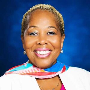 A person with short blond hair, white earrings, and a colorful scarf smiling against a blue background.