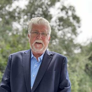 An older man with white hair, beard, and glasses is wearing a blue shirt and dark blazer. He is standing outdoors with a background of green, leafy trees.