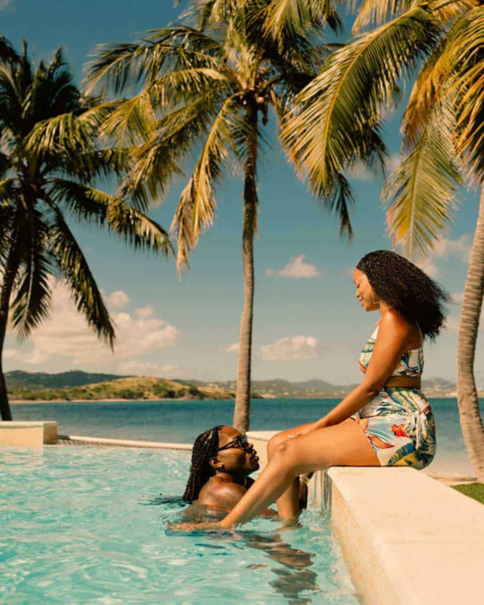 Two people enjoy a sunny day by a tropical pool. One is in the pool, leaning against the edge, while the other sits on the poolside. Palm trees and a clear sky are in the background.